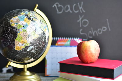 Close-up of apple over books by globe on table with text on blackboard in background