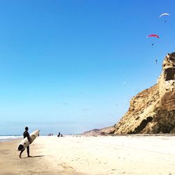 Side view of man with surfboard walking at beach against blue sky during sunny day