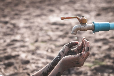 Cropped dirty hands of person below faucet on barren land during drought