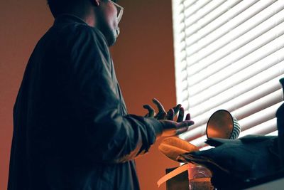 Midsection of man gesturing against blinds at home