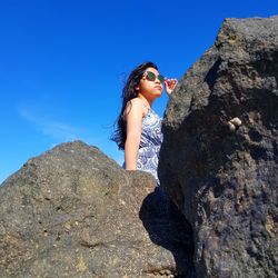 Side view of young woman standing on rock against blue sky