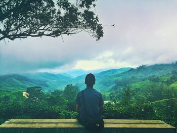 Rear view of man sitting on mountain against sky