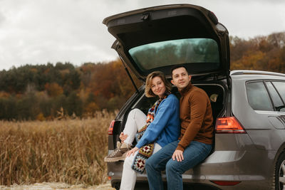 Happy young woman and man sitting in the open trunk of a car while traveling in autumn