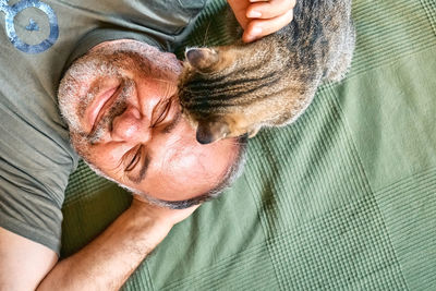 Top view of bearded middle-aged man lying on a bed and playing with his gray tabby cat.