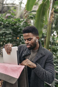 Young man putting laptop in bag against plants