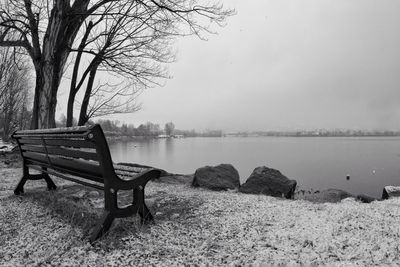 Empty bench by bare tree by lake against sky