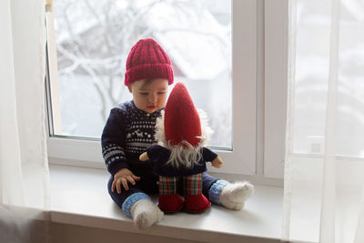 Baby boy sitting with soft toy gnome on the window
