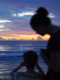 Silhouette woman with son standing at beach against sky during sunset