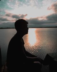Side view of man sitting by lake against sky during sunset