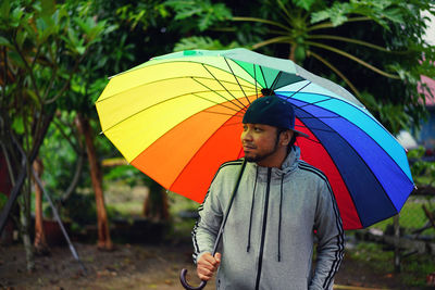 Man holding colorful umbrella while walking against trees during rainy season