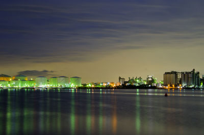 Illuminated factory building by sea against sky at night