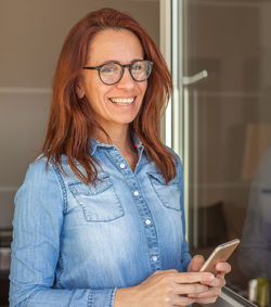 Mature woman using mobile phone while standing by window at home