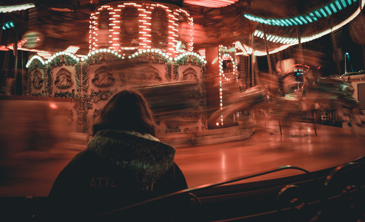 REAR VIEW OF WOMAN IN ILLUMINATED AMUSEMENT PARK