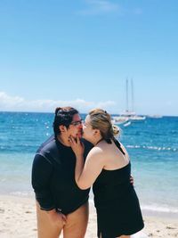 Lesbian couple kissing while standing at beach against blue sky