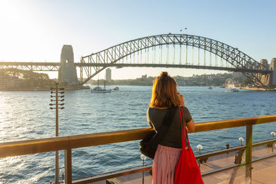 Rear view of woman looking at sydney harbor bridge over river against sky
