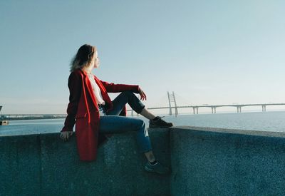 Side view of woman sitting on retaining wall by sea against clear sky