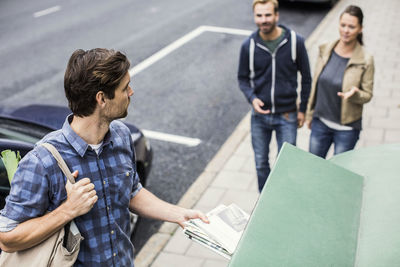 Man putting newspaper in recycling bin while looking at friends on sidewalk