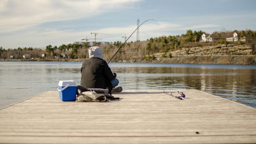 Rear view of man sitting on pier over river against sky