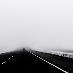 Car moving on road amidst snow covered field against sky in foggy weather