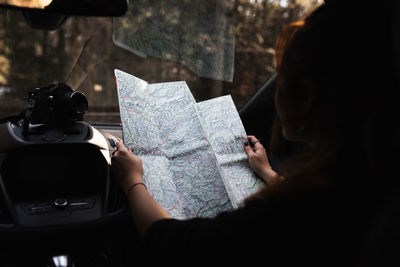 Unrecognizable female traveler sitting on passenger seat and reading tourist map while checking route during road trip through woodland