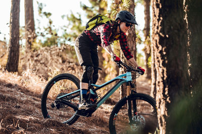 Side view of woman with backpack riding on bicycle on path between trees on hill in forest