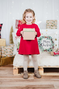 Happy little girl wearing red christmas dress at home over christmas decoration. holding gift box