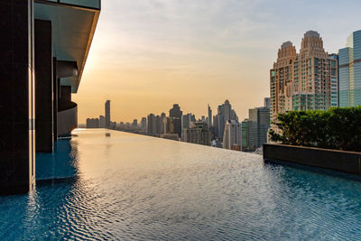 Infinity pool by modern buildings in city against sky during sunset