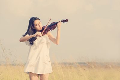Young woman playing violin while standing on field against sky