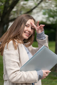 Portrait of smiling teenage girl holding books against trees