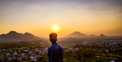 Rear view of man looking at cityscape against sky during sunset