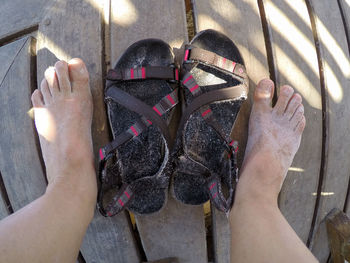 Low section of man standing by dirty sandals on wooden plank