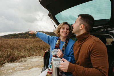Happy young woman and man sitting in open trunk of car while traveling in autumn, road trip concept