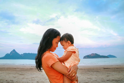 Mother holding baby girl while standing at beach against sky