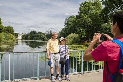 Son photographing parents with mobile phone standing on footbridge over river against sky at park during sunny day