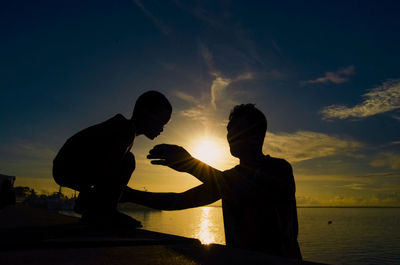 Silhouette father and son standing by sea against sky during sunset