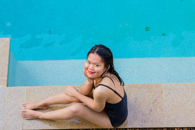Beautiful asian woman sitting at pool side on sunny day while smiling