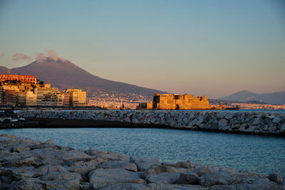 Buildings by sea against sky during sunset in city