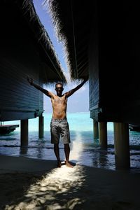 Shirtless man standing amidst stilt house at beach