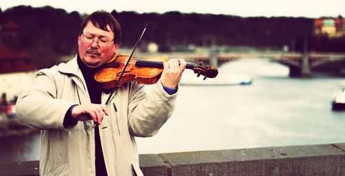 Mature man playing violin while standing on bridge against river