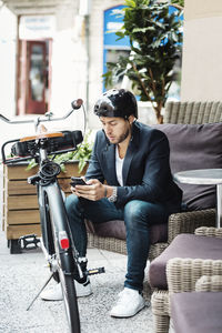 Businessman with bicycle using smart phone while sitting on couch at sidewalk cafe