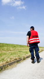 Rear view of firefighter walking on road by grass field against sky