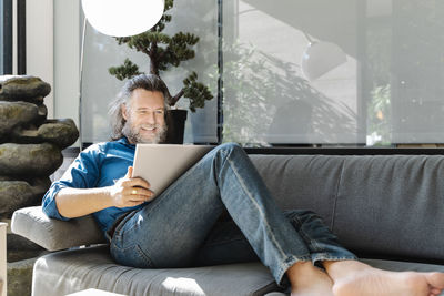 Mature man with a beard smiling and working with his laptop on a sofa at home. business concept