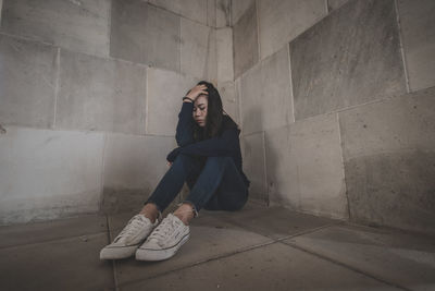 Young woman looking away while sitting on floor against wall