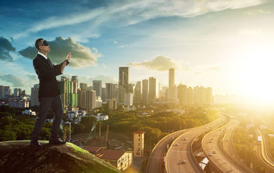 Full length of businessman with blindfold standing on cliff in city during sunset