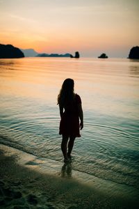 Rear view full length of woman standing on shore at beach during sunset