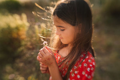 Side view of adorable dreamy girl in red dress standing with dandelion on meadow in bright sunny day and