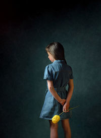 Back view of a young girl long brown hair blue dress standing holding yellow tulip behind her back