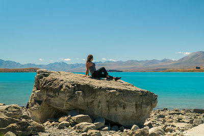 Man sitting on rock by sea against blue sky