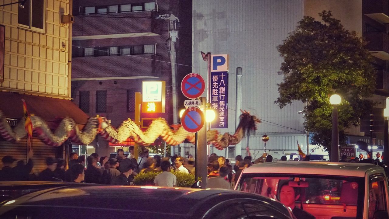 city, building exterior, architecture, men, built structure, large group of people, person, illuminated, street, text, lifestyles, communication, city life, transportation, leisure activity, night, land vehicle, tree, outdoors