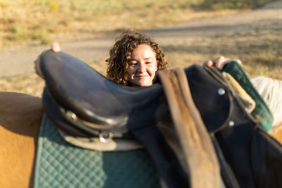 Middle aged female with curly hair putting saddle on back of mare on farm in sunlight